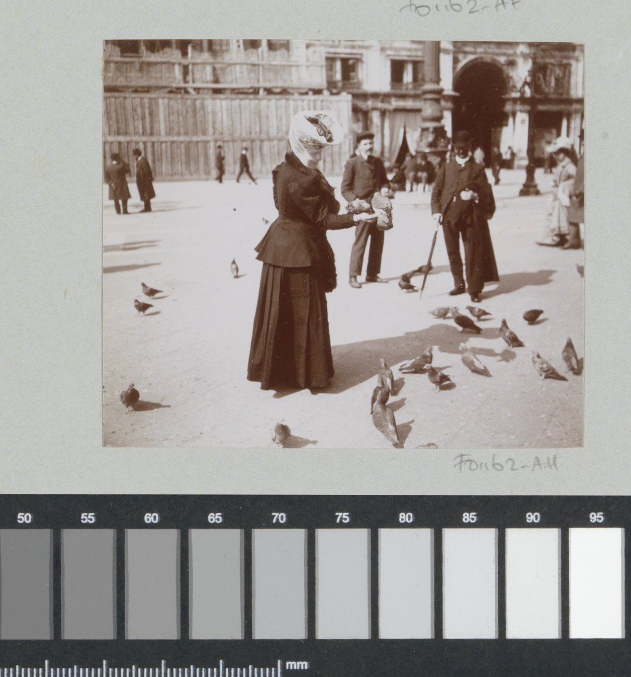 Black and white griny picture of an elegantly dressed woman feeding pigeons, two men standing a bit further watching her